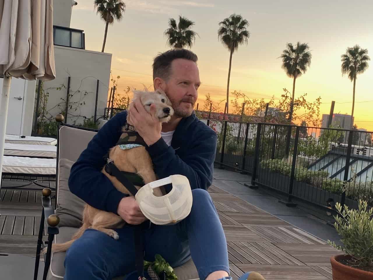 Jason-gray stanford holding his dog with view on sunset and palm trees in the background.