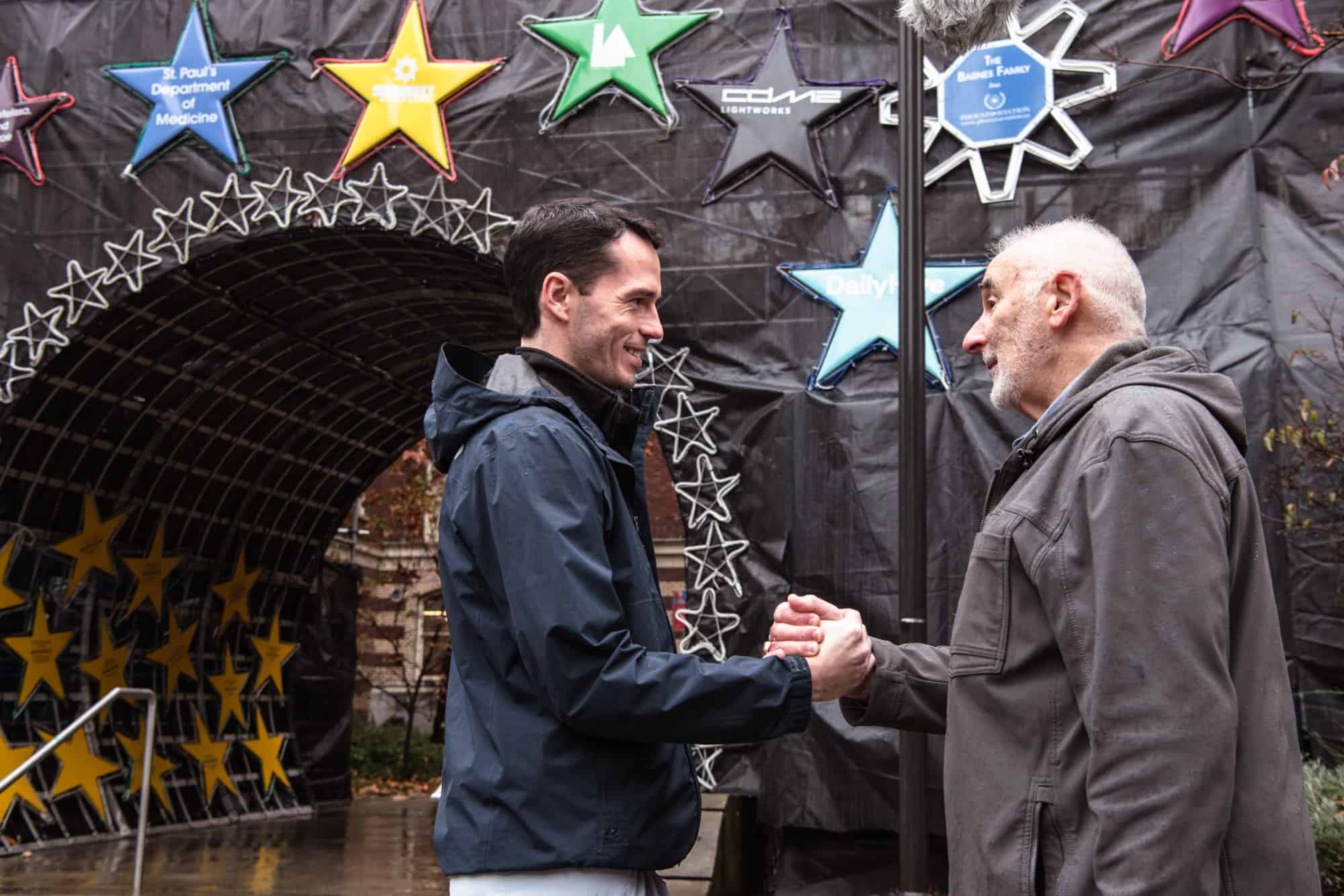 A photo of a nurse at St. Paul's Hospital, Luke Currie, and grateful patient, James Stitchman, shaking hands while standing outside St. Paul's Hospital in front of the Lights of Hope star display.