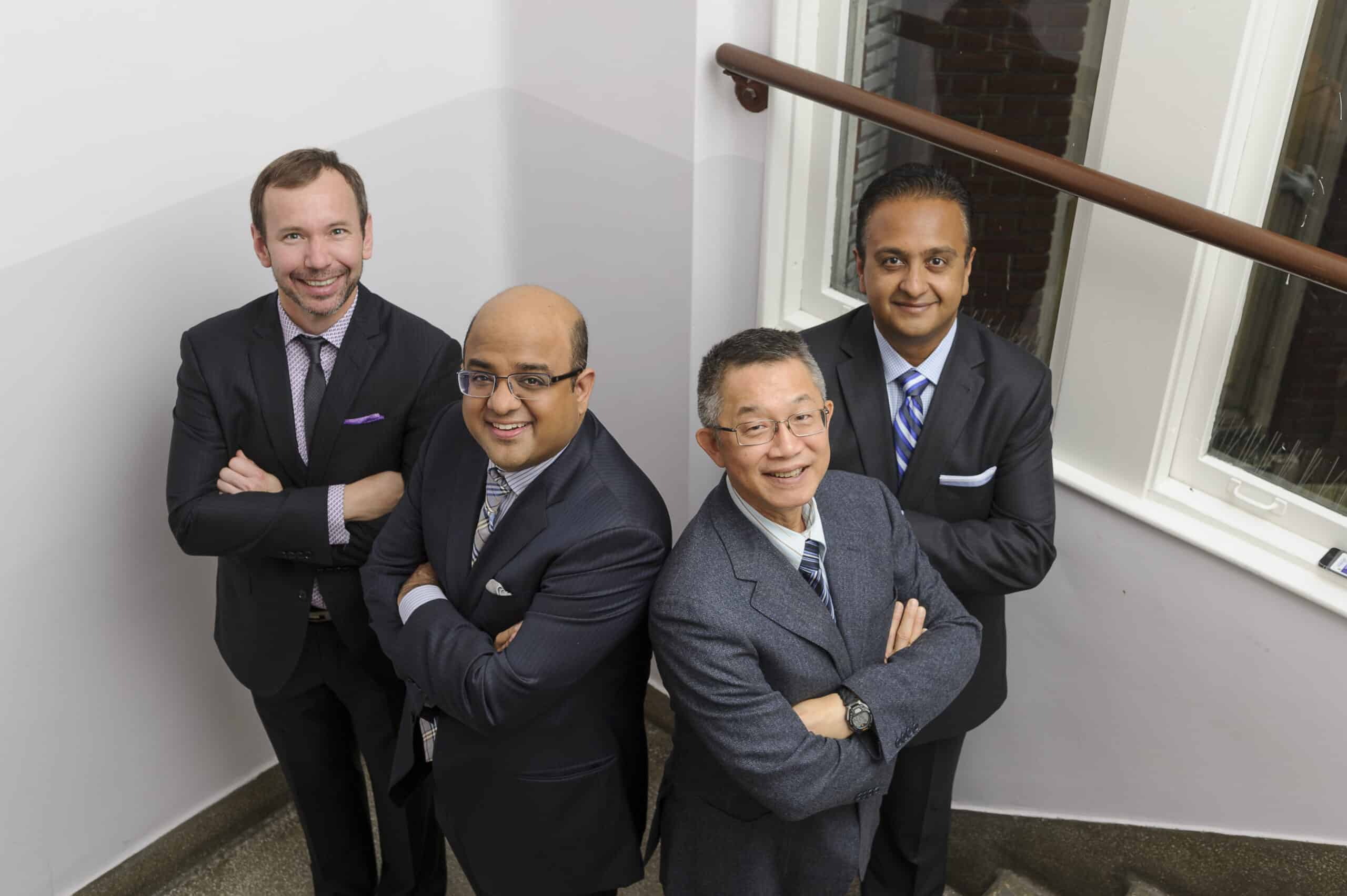 Four doctors, in suits with arms crossed on a stairwell, look up with smiles at the camera.