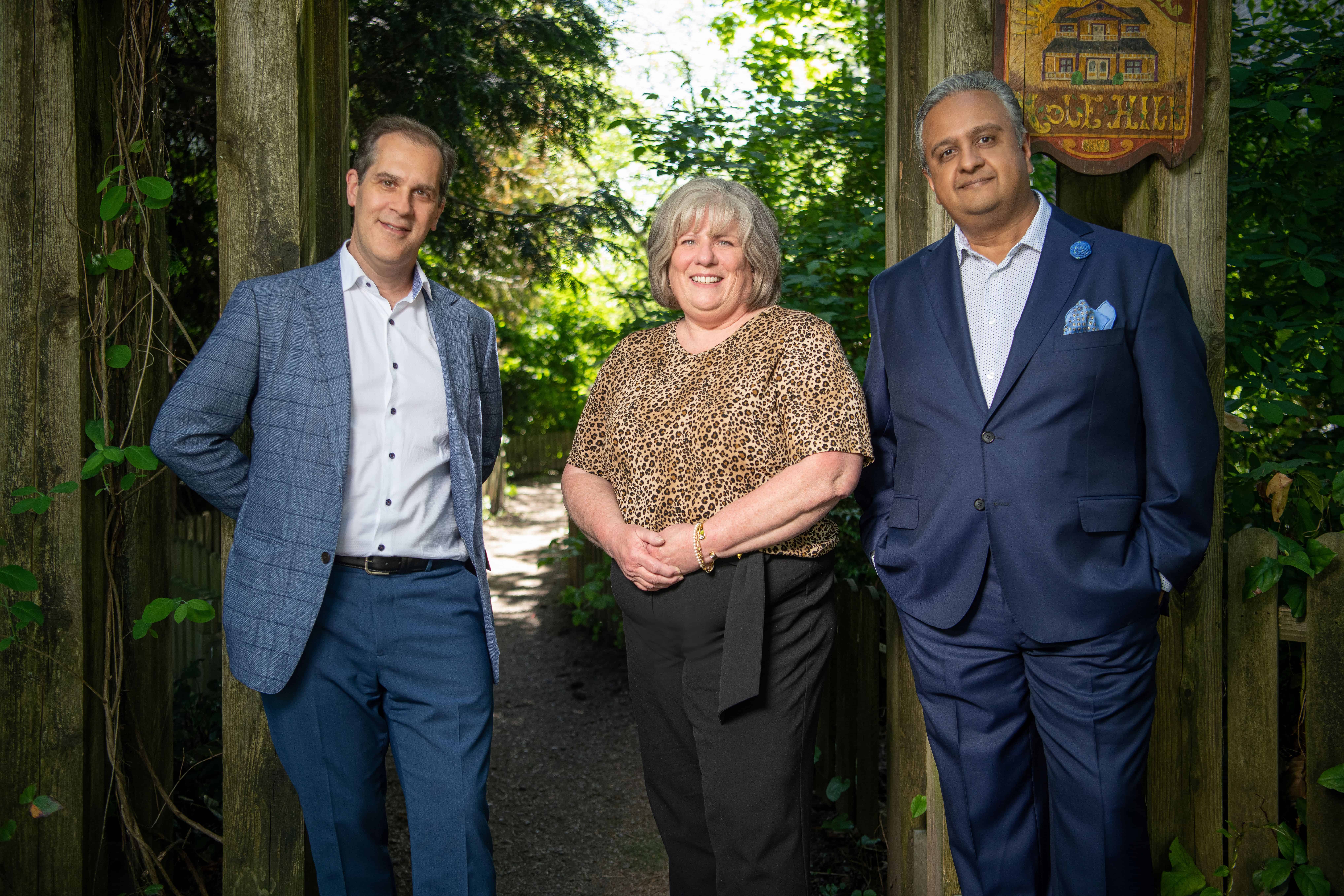 Dr. Bressler and Dr. Raval, wearing suits, stand on either side of Patti amid the trees.