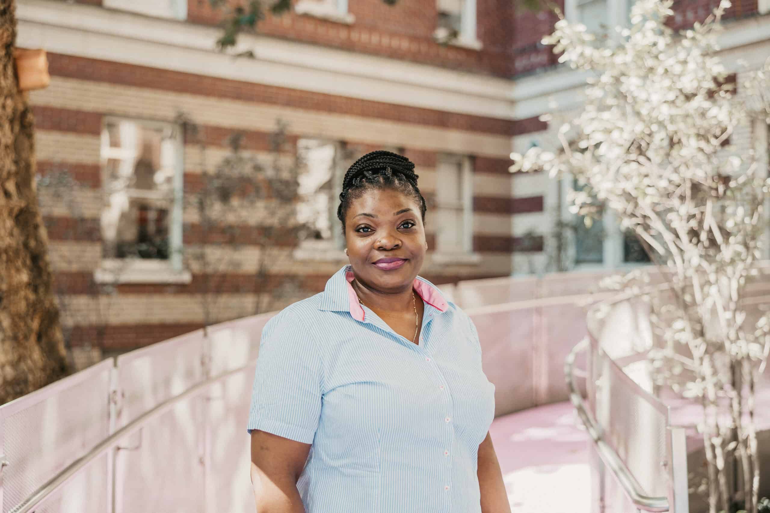 Ajoke, wearing a blue and white striped shirt with a pink collar, stands on the ramp outside St. Paul's Hospital