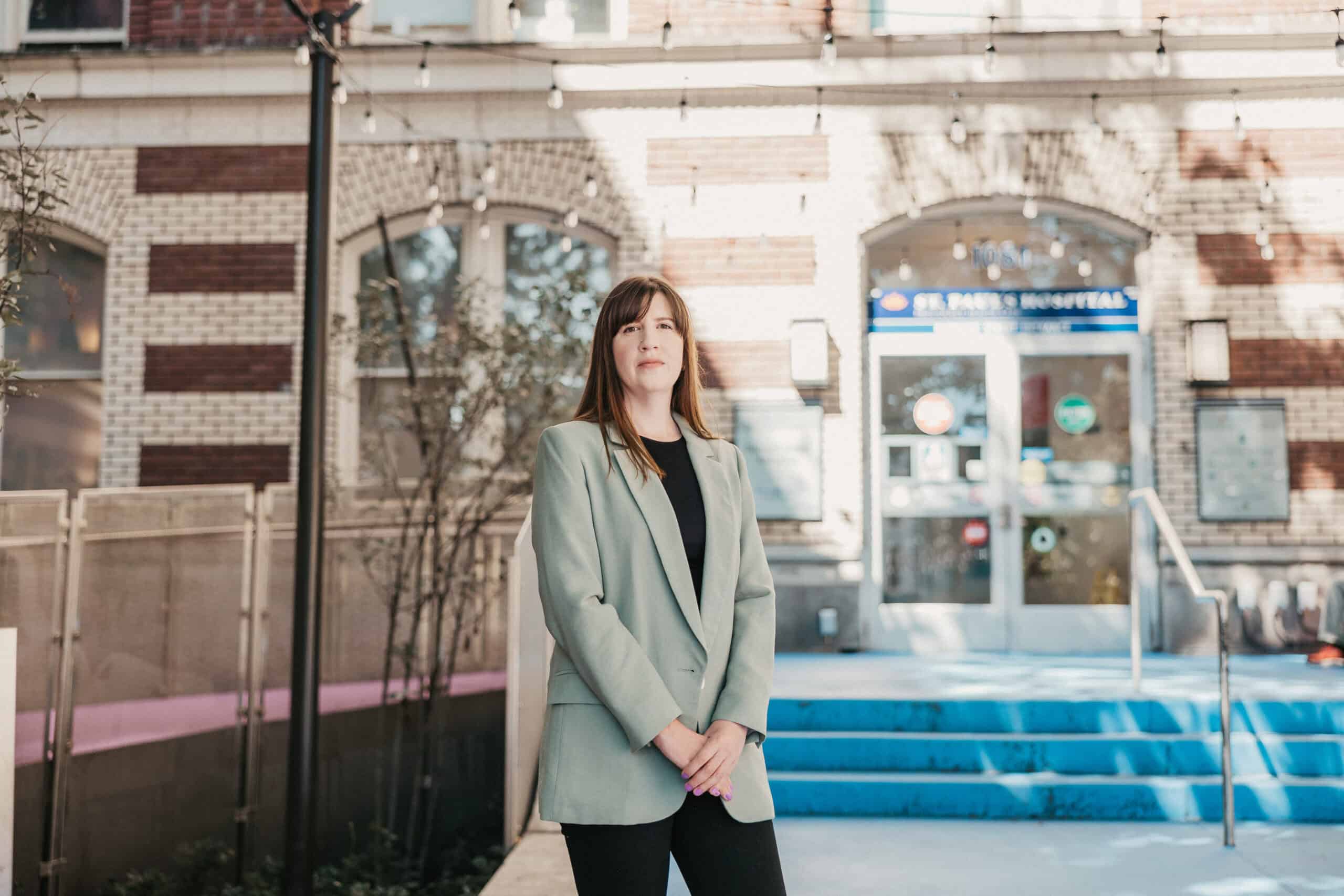 Clare Carrigan, psychologist, stands with her hands clasped in front of her in front of the doors of St. Paul's Hospital.