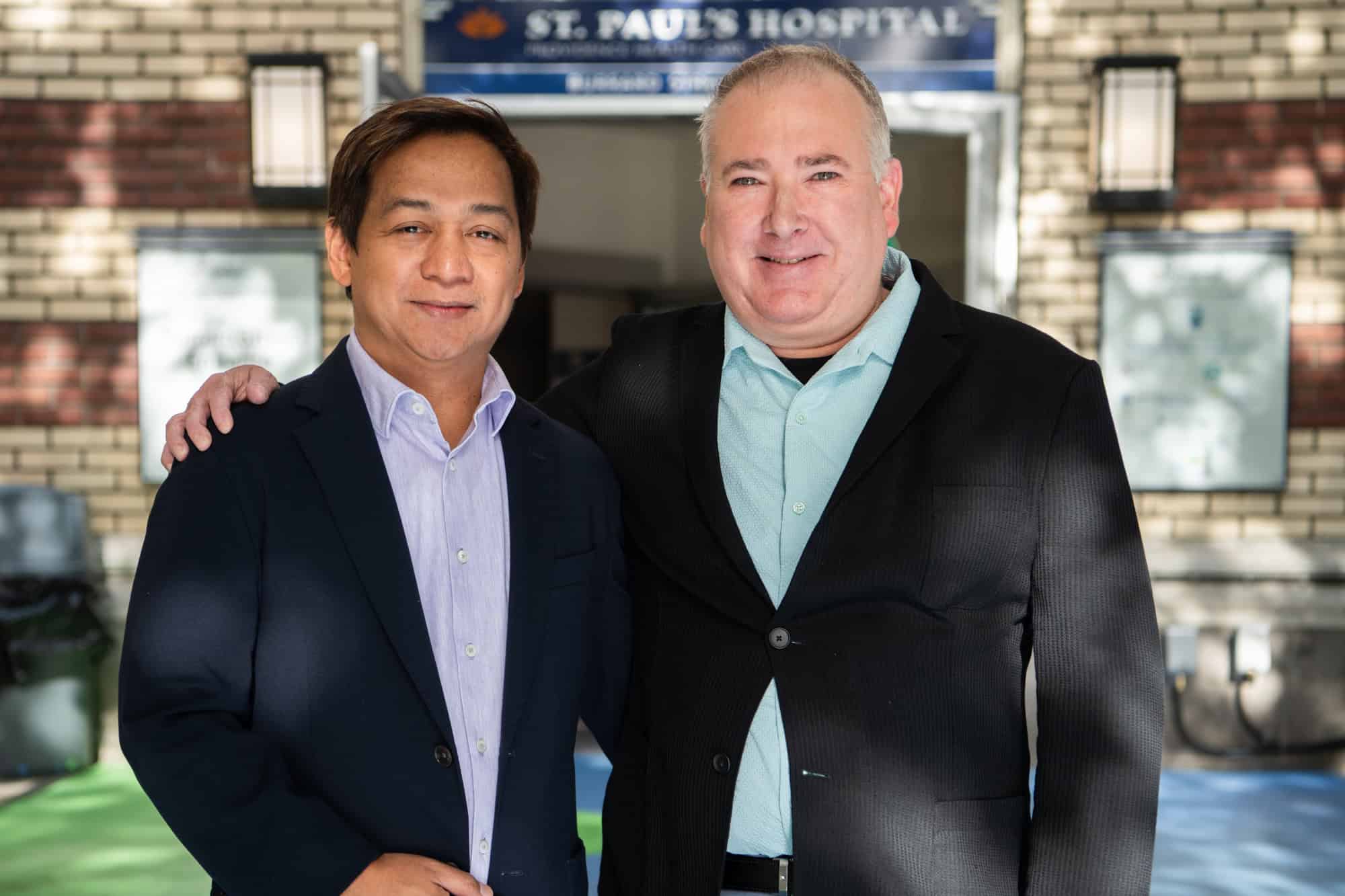 Two men in suits are smiling in front of St. Paul's Hospital.
