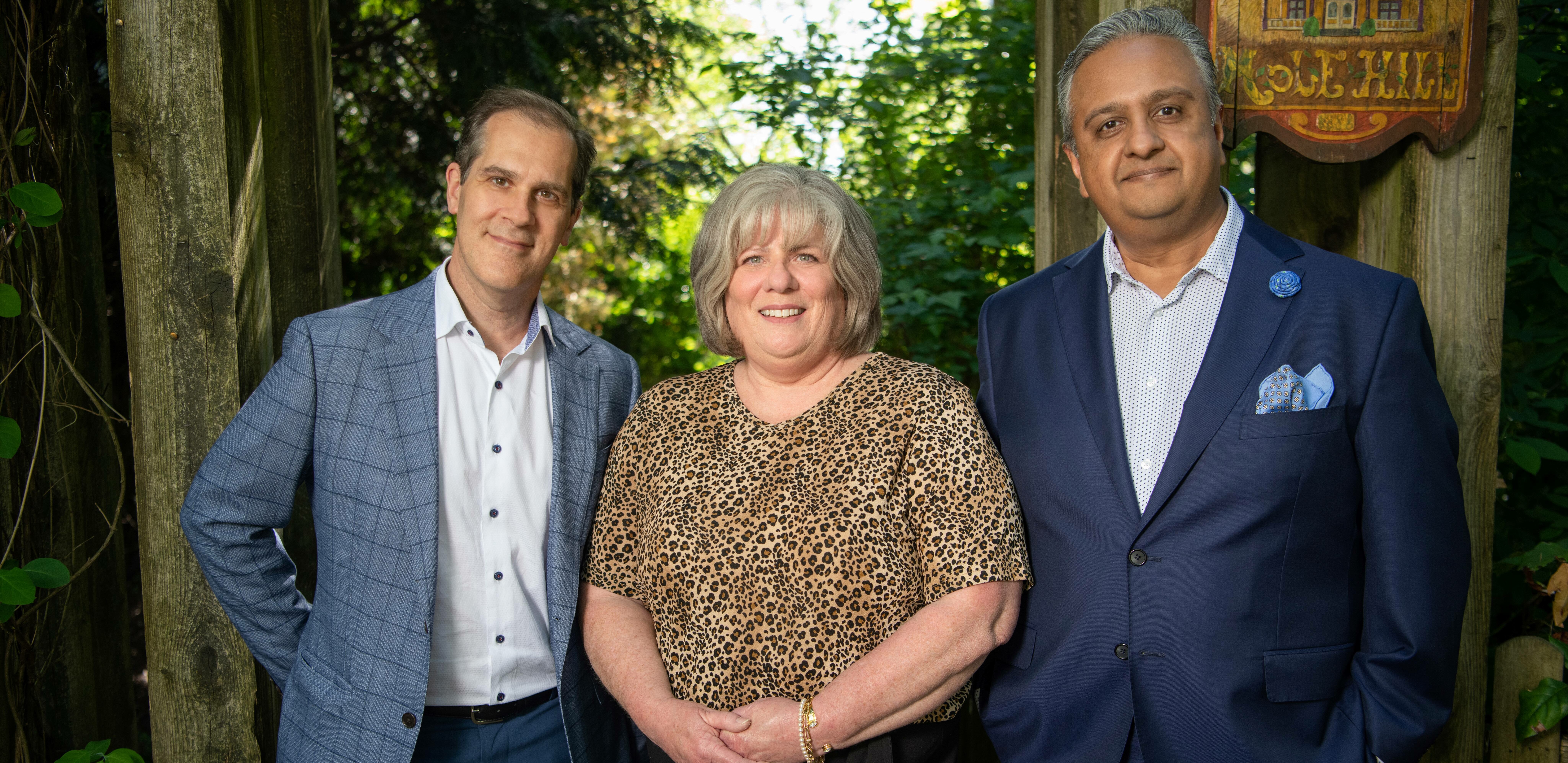 Dr. Bressler and Dr. Raval, wearing suits, stand on either side of Patti amid the trees.