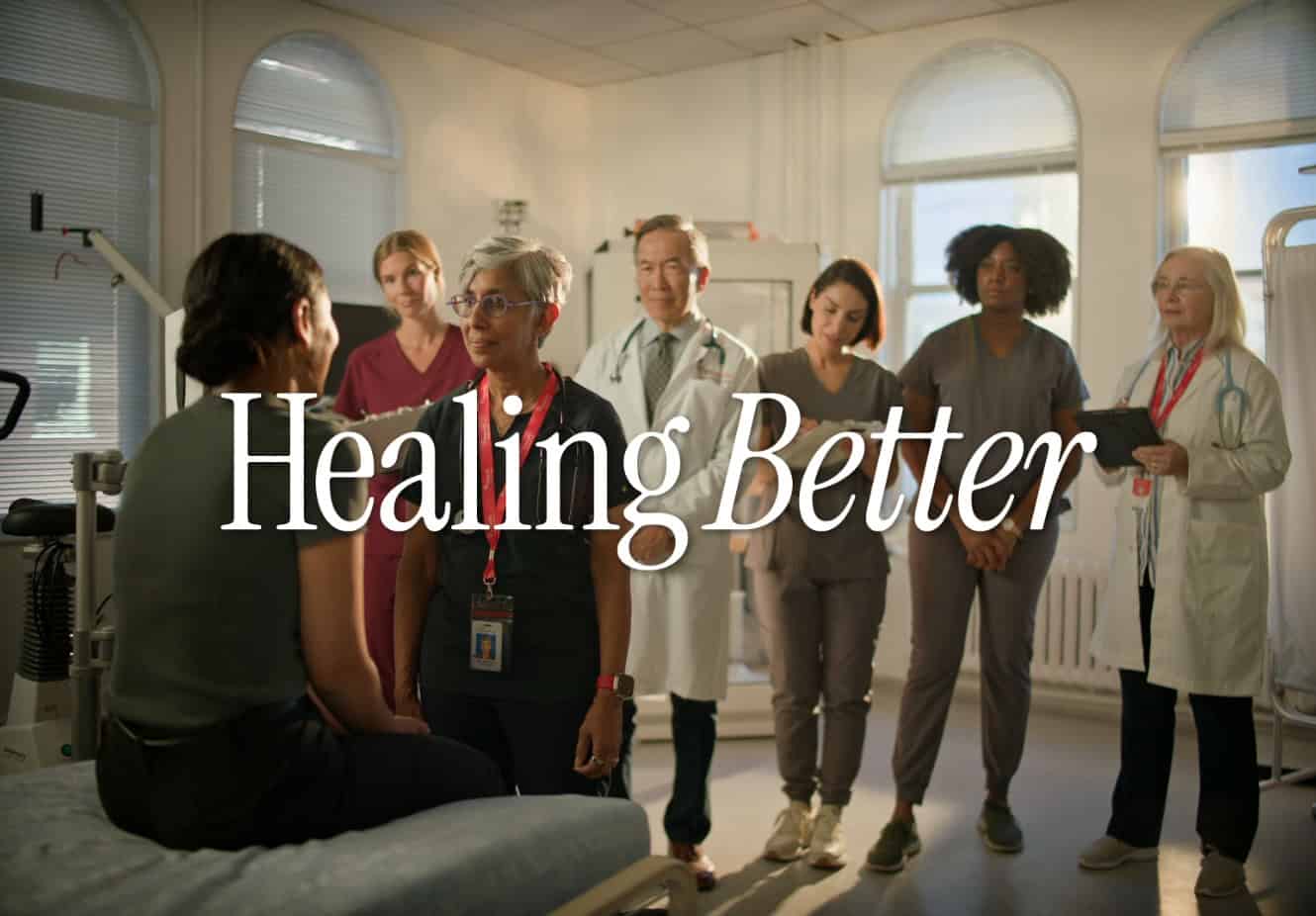 Doctor at St. Paul's Hospital listening and standing in front of a patient sitting on bed, with other doctors and health care professionals standing behind them. With Healing Better text overlay.