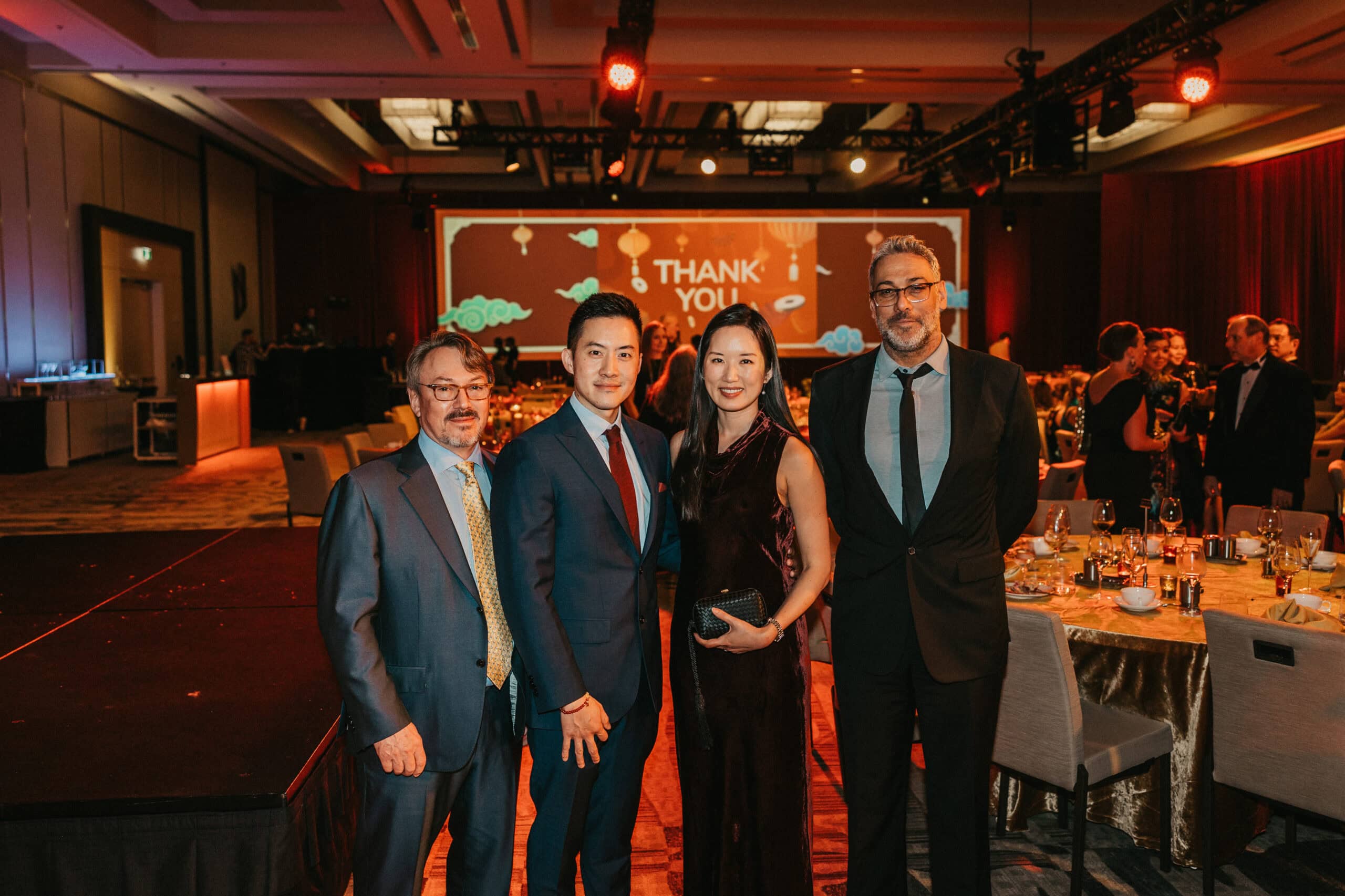 Four people in formal attire pose together at a gala event with a decorated stage and tables in the background.