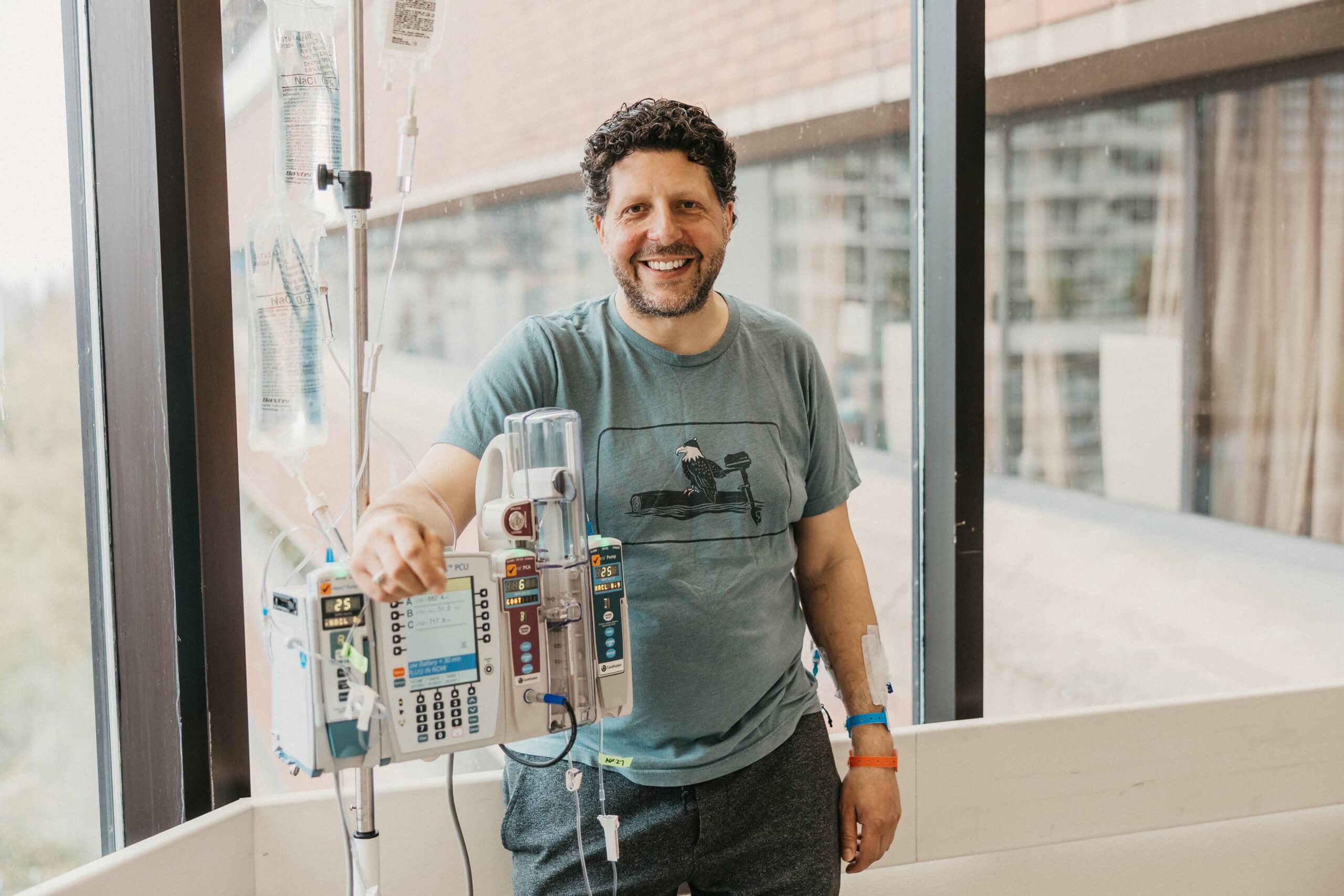 Angelo stands in the hallway at St. Paul's Hospital, in front of a window, hand on his IV stand. He is smiling.