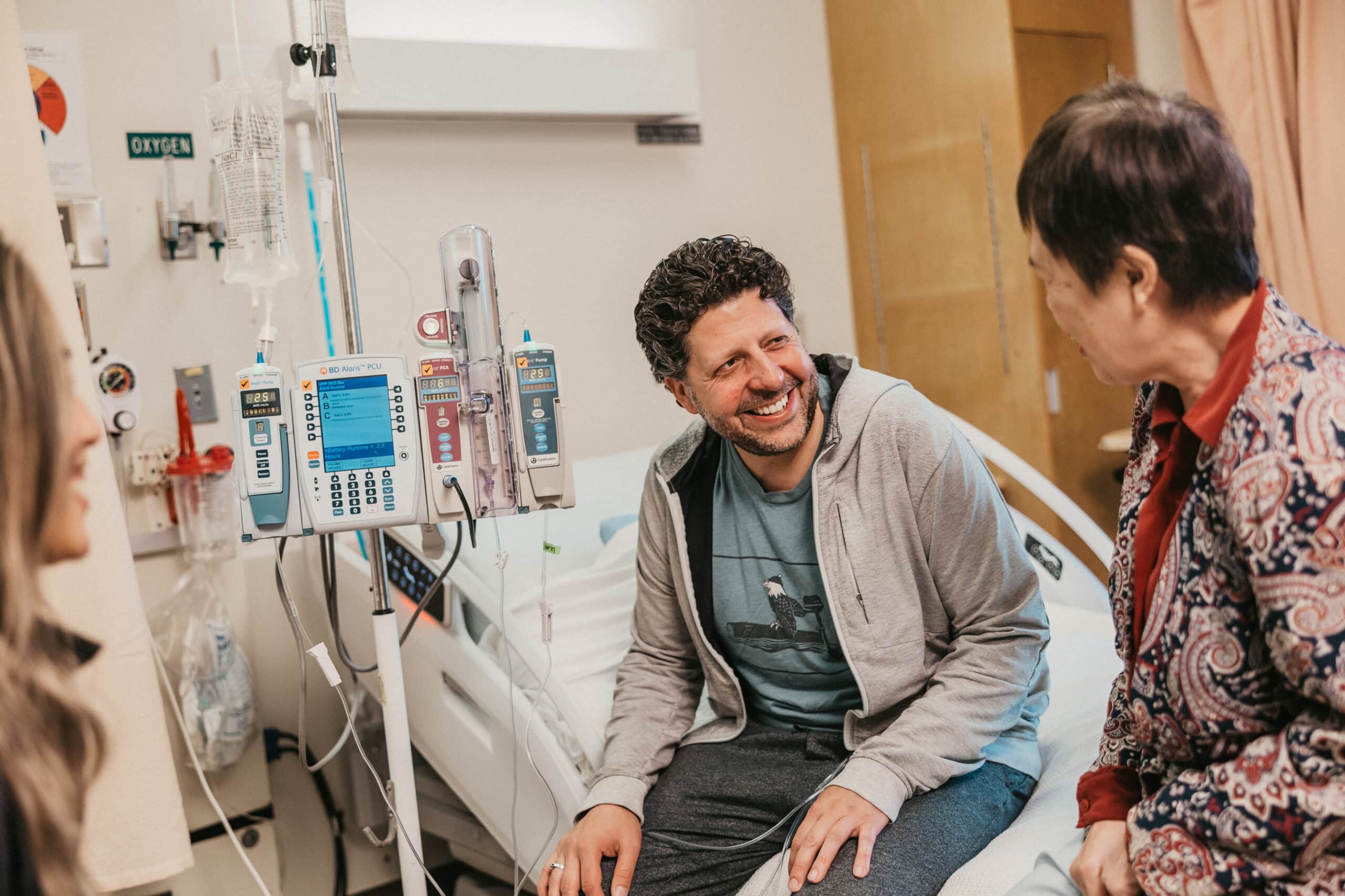 Patient Angelo sits on a hospital bed, attached to an IV, while smiling at his doctor - Dr. May Ong.