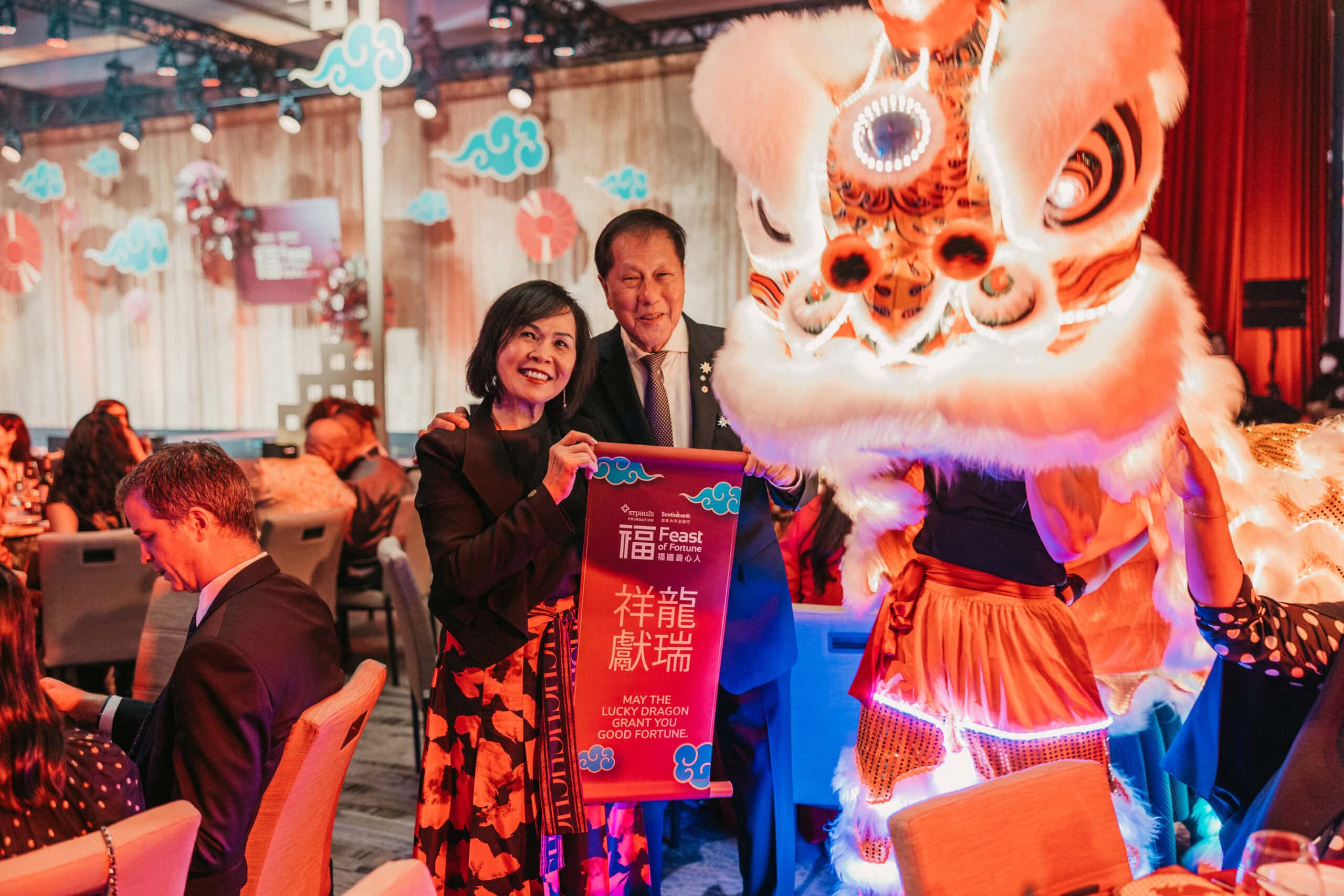 Patricia and Sing Yeo stand, smiling, alongside a lion dancer for Lunar New Year.