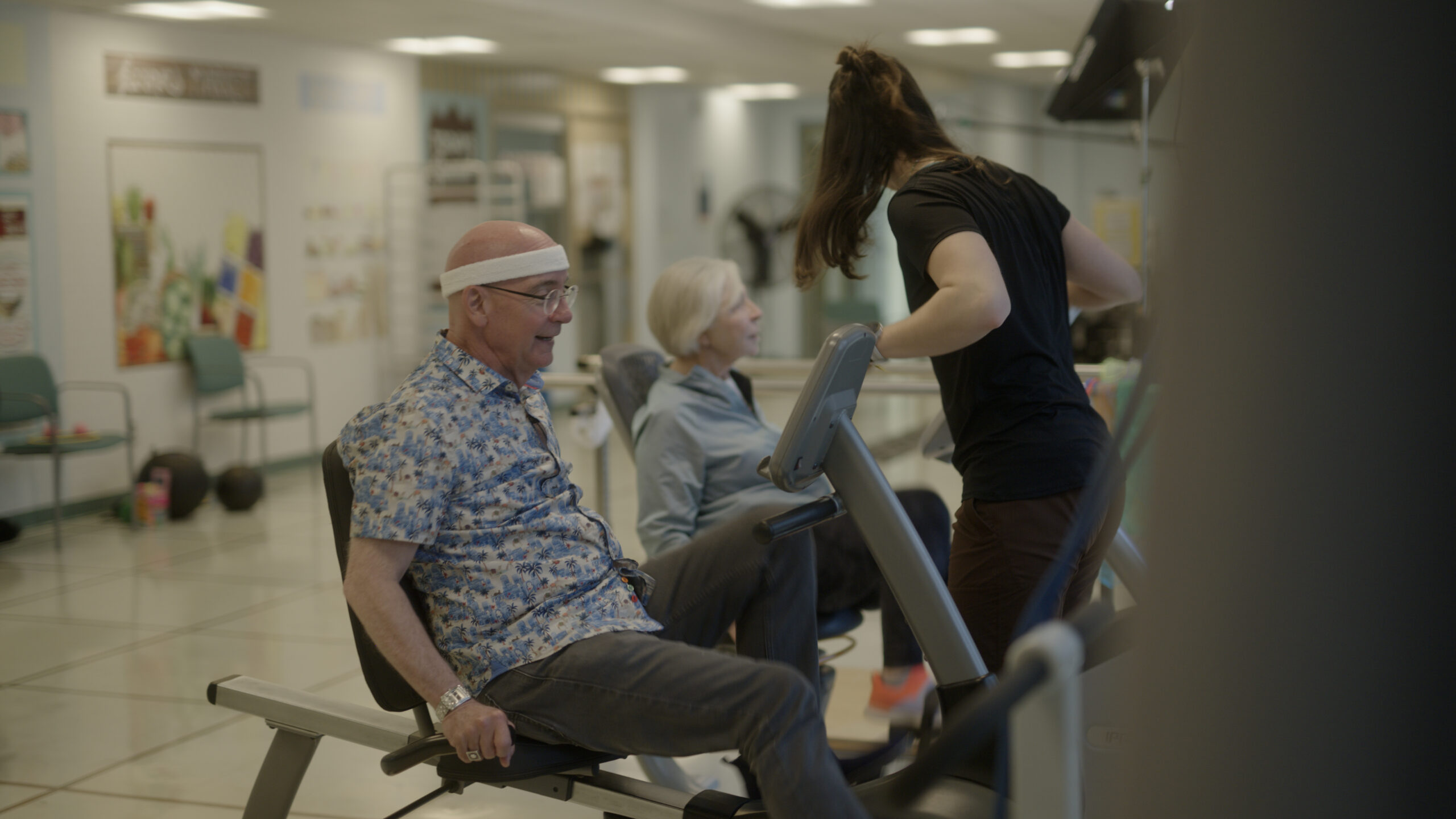Two older adults sit on recumbent bikes in the gym.