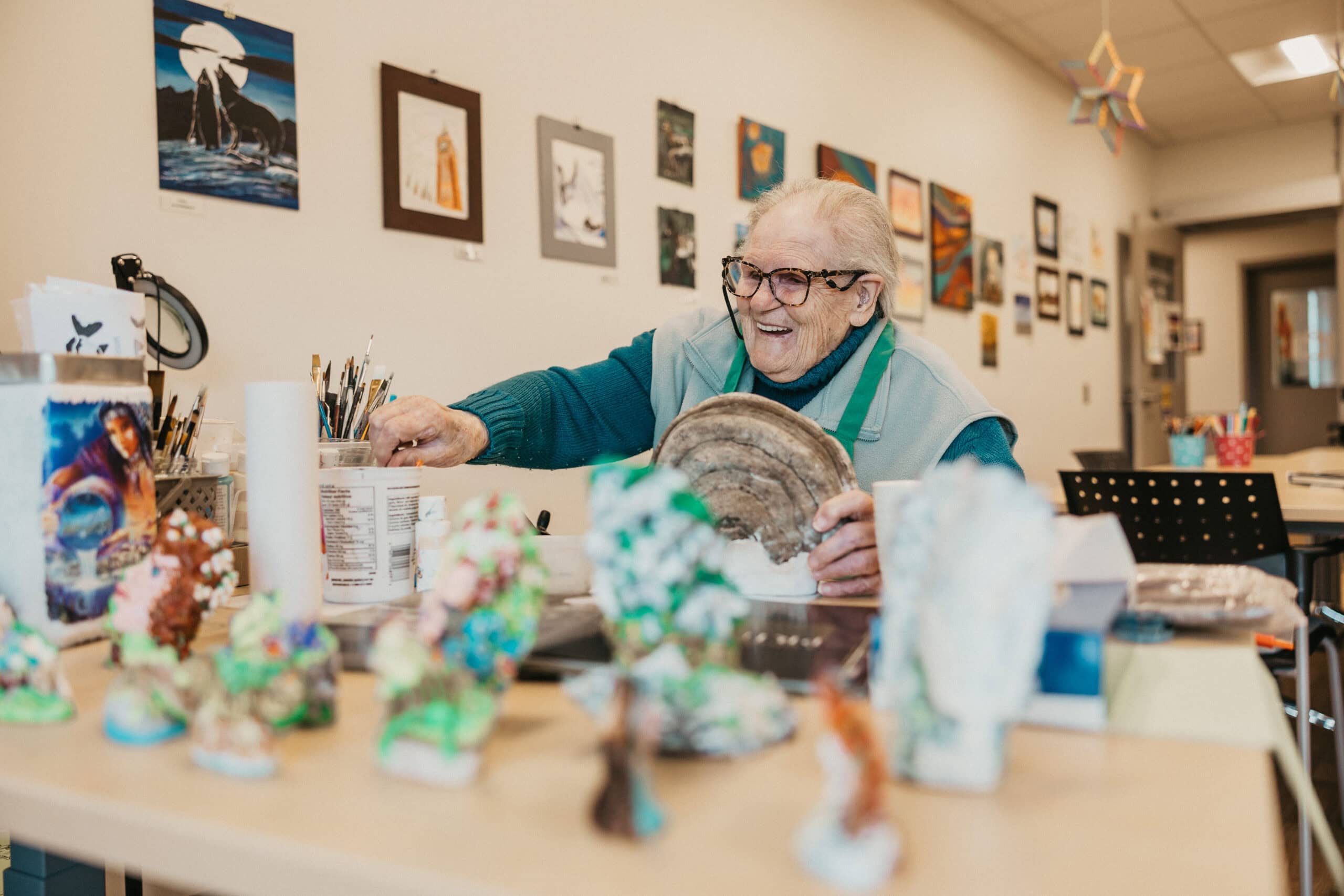 An older adult smiles as she sits painting pottery in a bright art room.